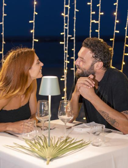 Cena romantica sulla spiaggia con luci soffuse, coppia felice e sorridente.