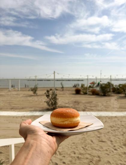 Mano offre una ciambella su spiaggia soleggiata, cielo azzurro e mare calmo.