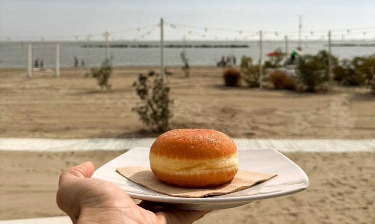Mano offre una ciambella su spiaggia soleggiata, cielo azzurro e mare calmo.