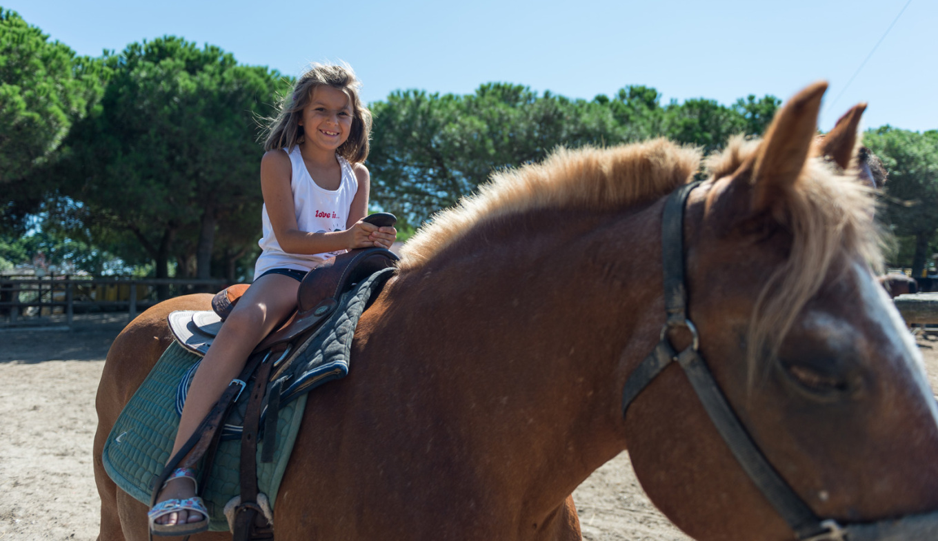 Bambina sorridente cavalca un cavallo al sole, circondata da alberi verdi.