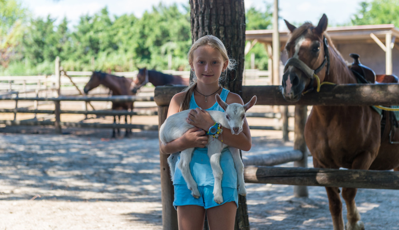 Bambina felice abbraccia un capretto, circondata da cavalli in un ranch soleggiato.