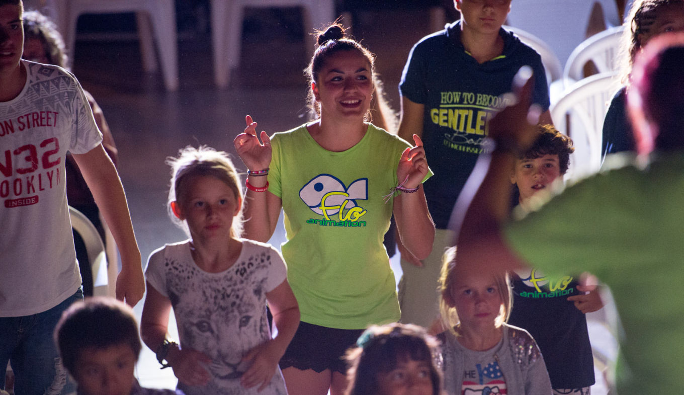 Bambini e animatrice sorridenti, atmosfera gioiosa e vivace, luci soffuse, momenti di festa.