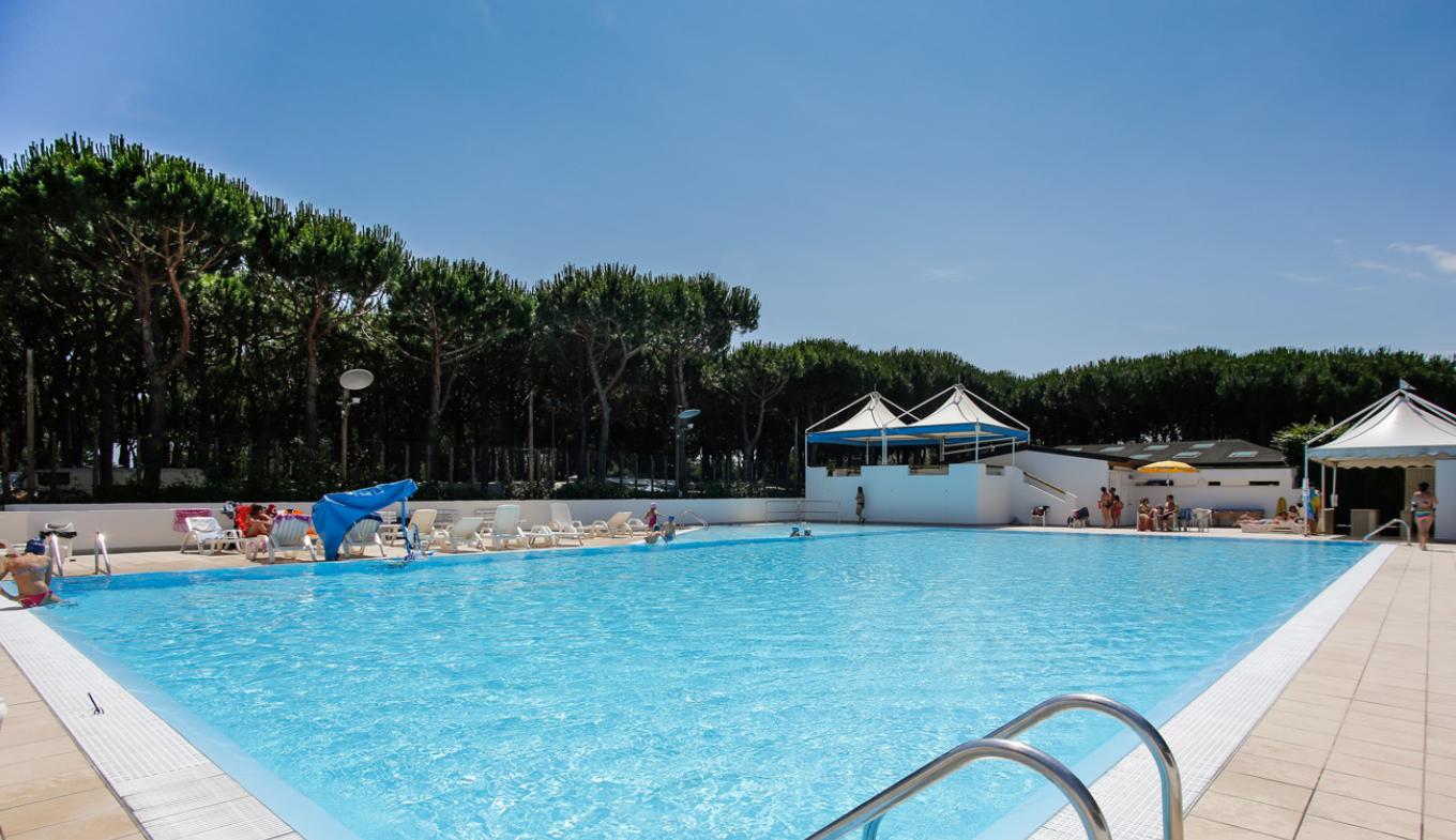 Piscina all'aperto con lettini, cielo azzurro, atmosfera rilassante e alberi verdi sullo sfondo.