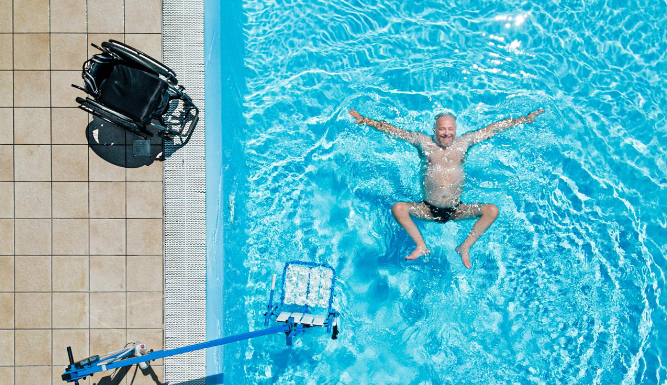 Un uomo felice galleggia in piscina, accanto a una sedia a rotelle.