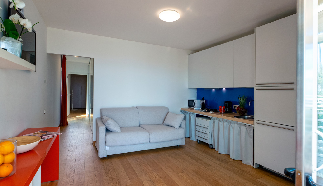 Helles, modernes Wohnzimmer mit grauem Sofa, weißer Küche und bunten Akzenten.