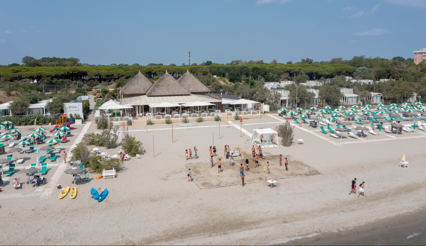 Spiaggia vivace con ombrelloni colorati, bambini giocano a pallavolo, cielo sereno e mare calmo.