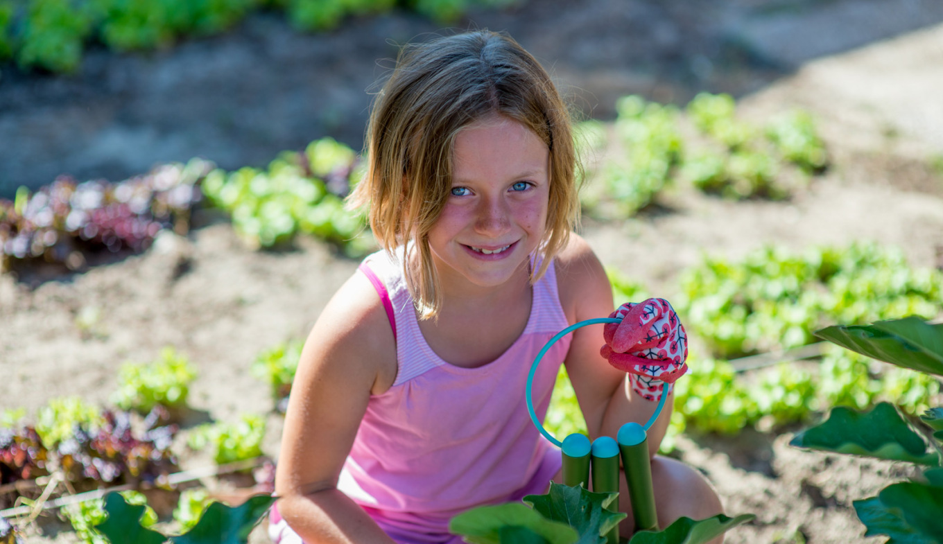 Bambina sorridente in giardino, guanti colorati, gioia e natura rigogliosa intorno.