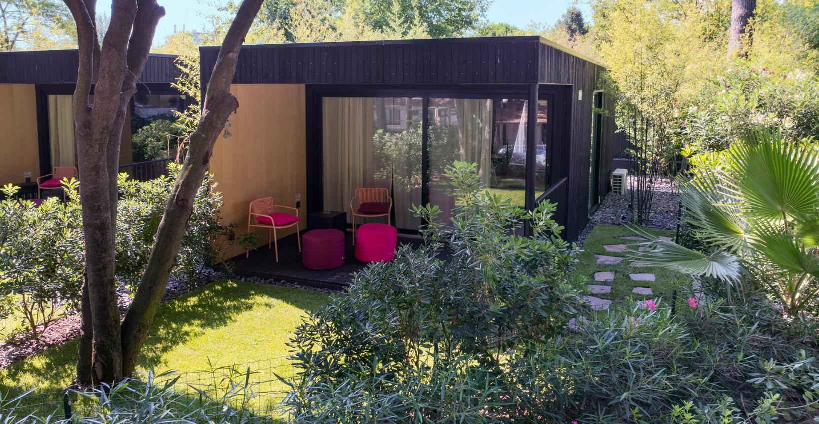 Giardino sereno con bungalow moderno, sedie colorate e natura rigogliosa avvolta dal sole.