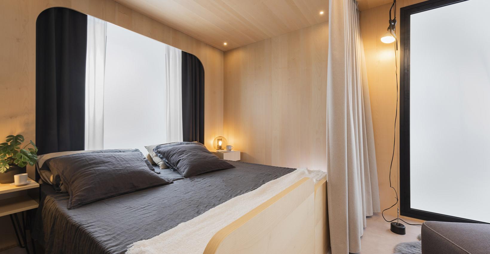 Camera minimalista, legno chiaro, letto accogliente, luci soffuse, atmosfera calda e rilassante.