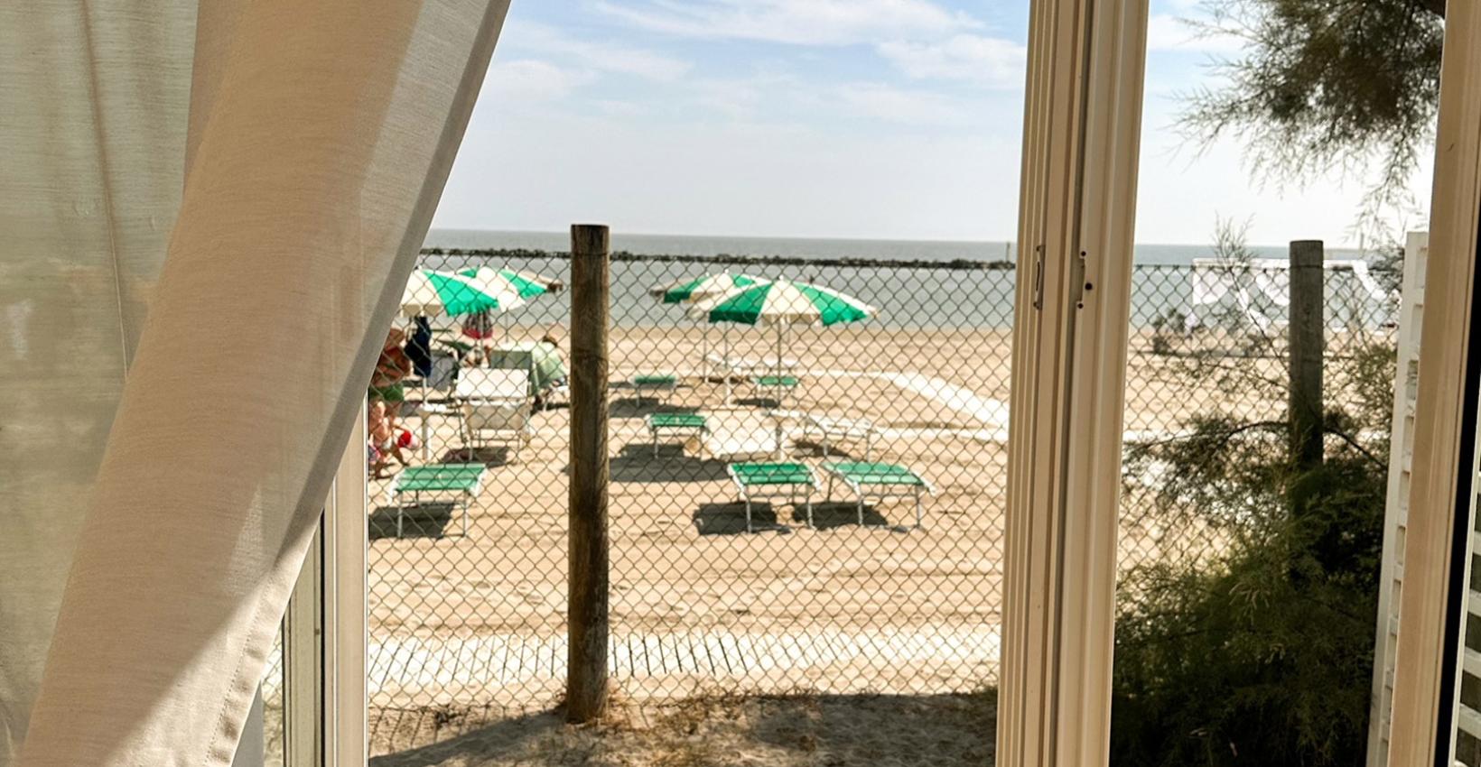 Vista mare da una veranda, tende mosse dal vento, ombrelloni verdi sulla spiaggia.