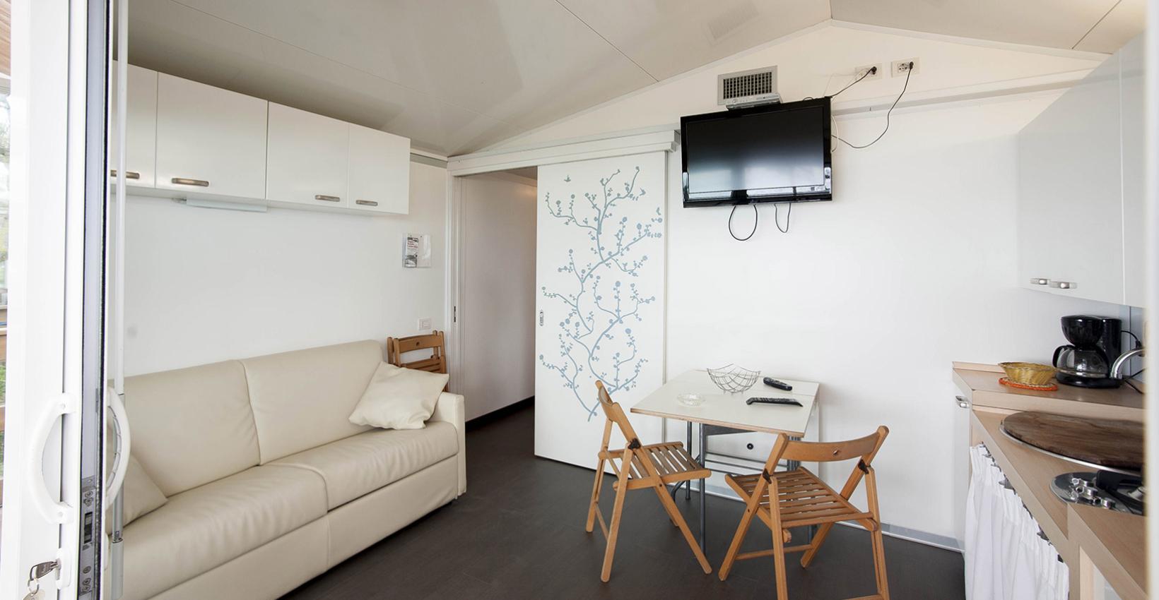 Interno moderno e luminoso, con divano bianco, tavolo, sedie e televisore.