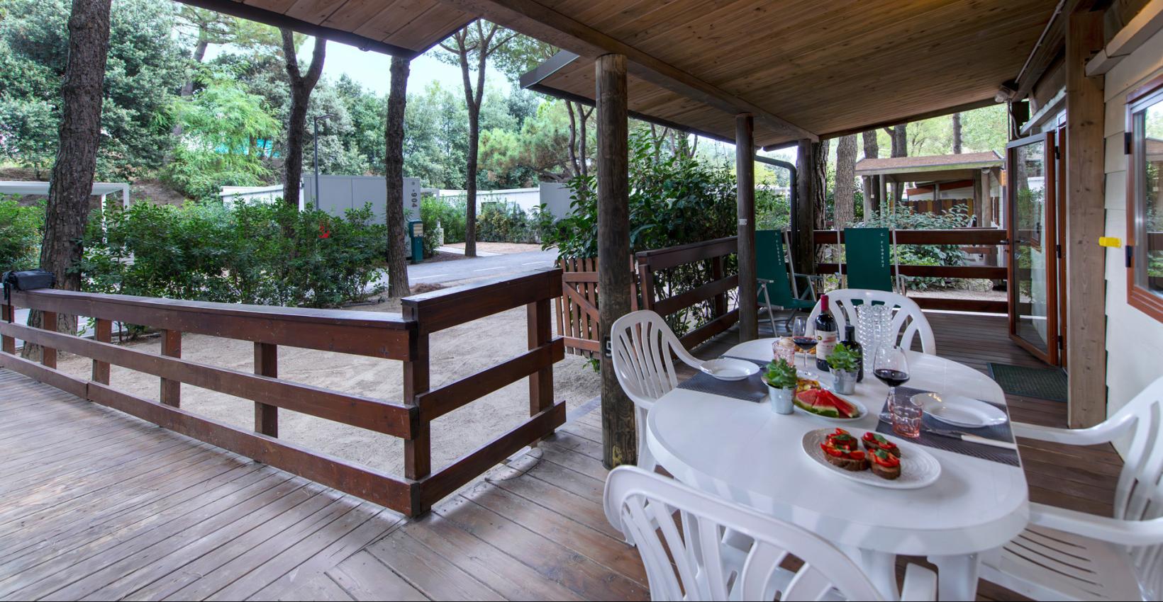 Terrazza in legno tra alberi, tavolo apparecchiato con vino e bruschette, atmosfera rilassante.