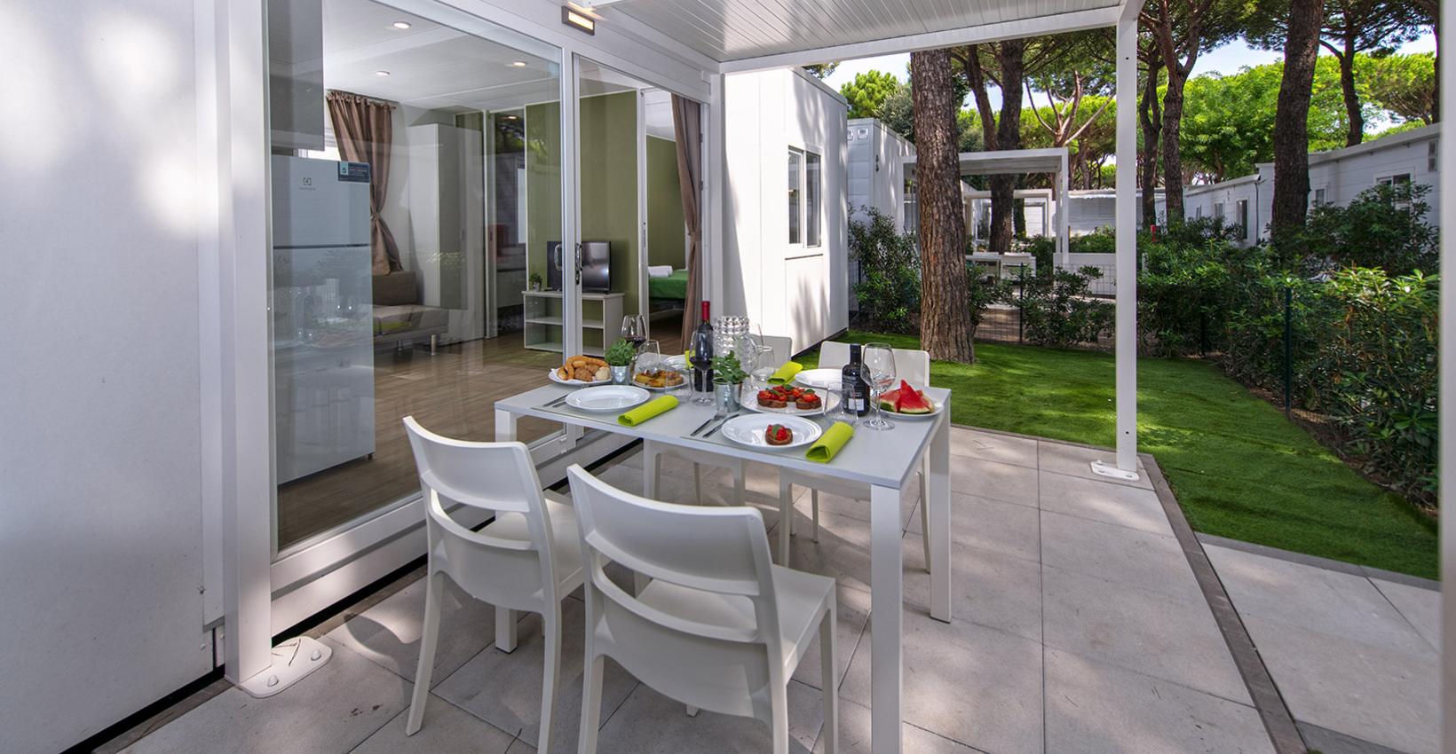Veranda luminosa con tavola apparecchiata, circondata da pini e verde rigoglioso.