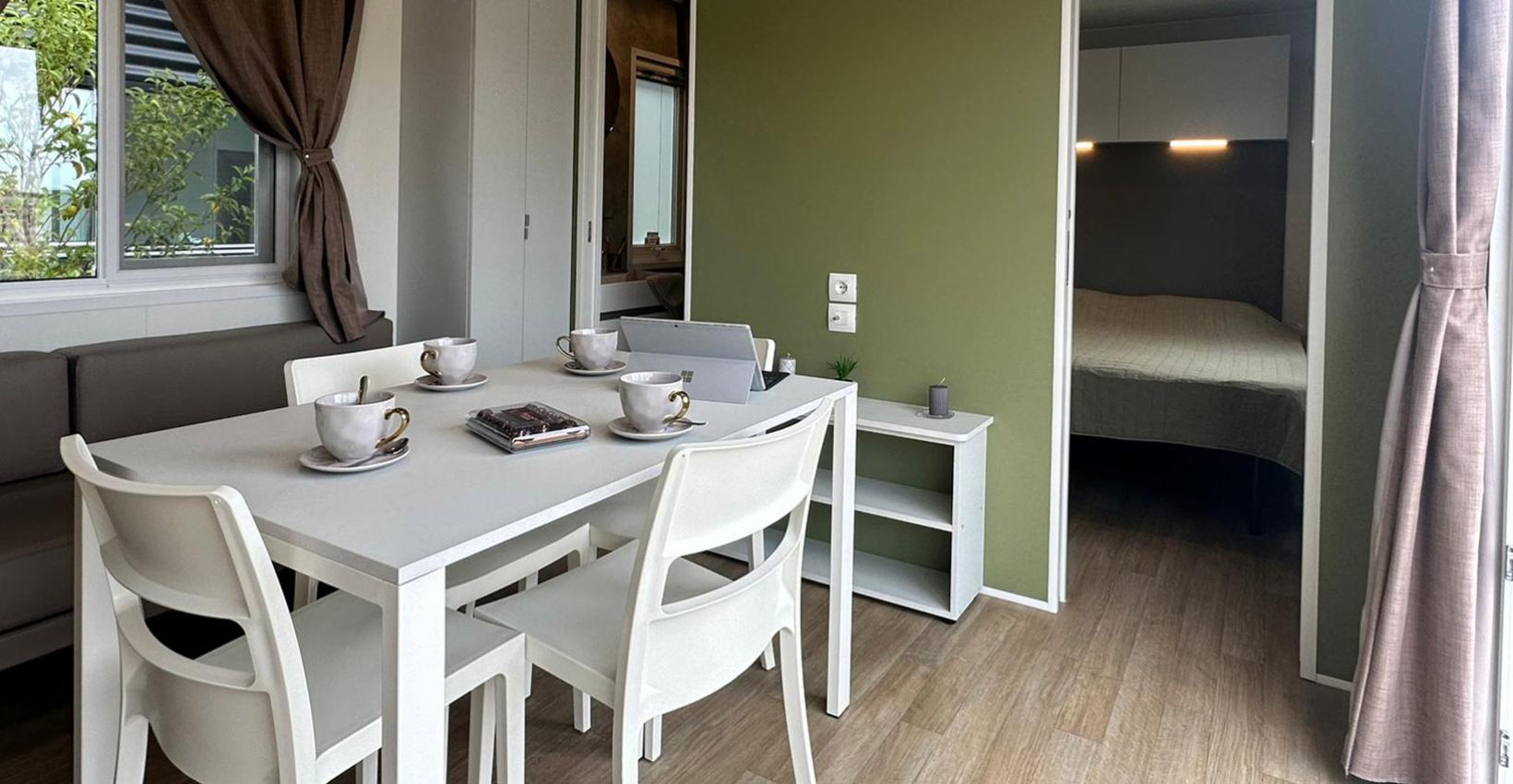 Interno accogliente con tavolo bianco, tazze eleganti e camera da letto visibile, atmosfera rilassante.