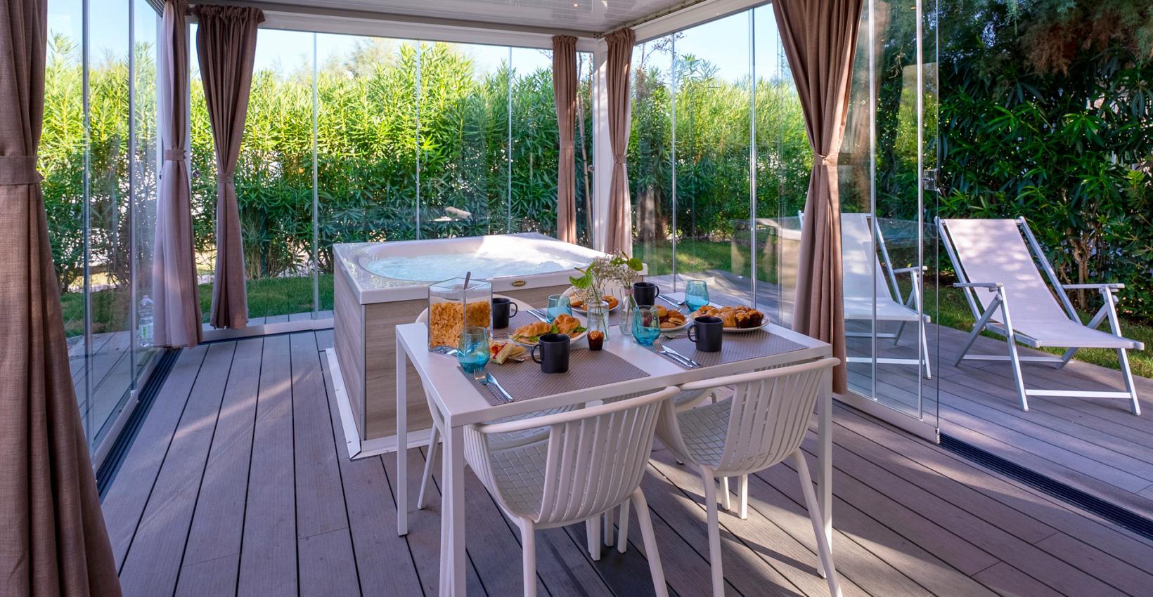Veranda luminosa con tavolo apparecchiato, vasca idromassaggio e vista su giardino verde rigoglioso.