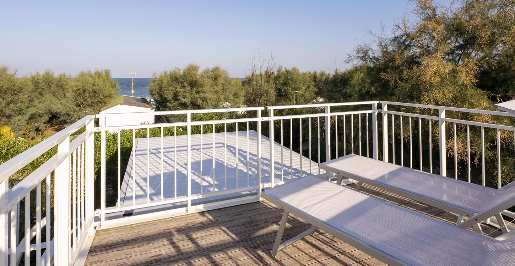 Terrazza soleggiata con vista mare, lettini bianchi, circondata da alberi verdi e cielo azzurro.