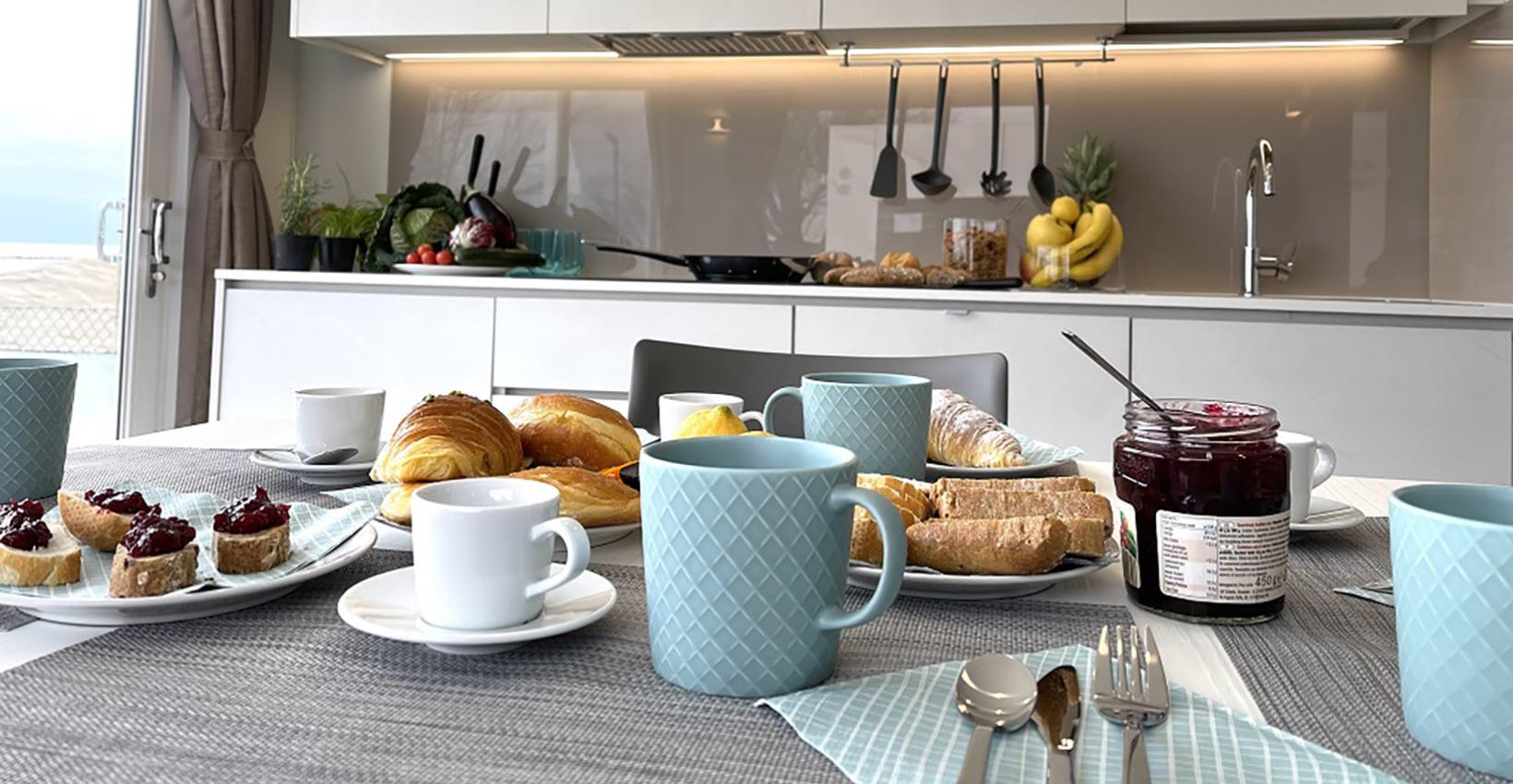 Colazione in cucina moderna: tazze azzurre, croissant, pane e marmellata, atmosfera accogliente e luminosa.