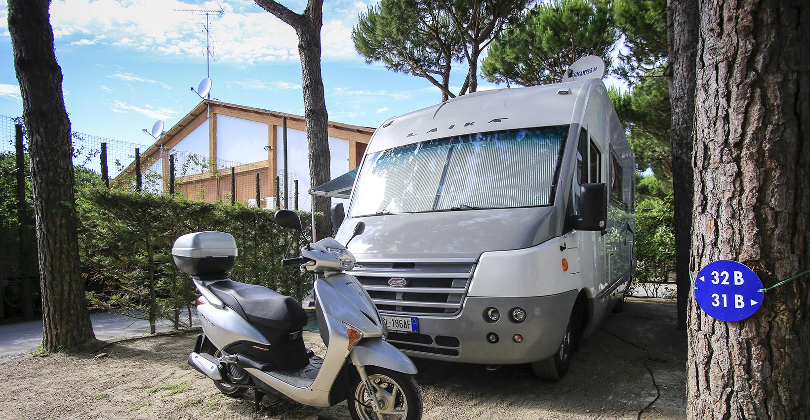 Camper e scooter in campeggio tra alberi, atmosfera rilassante e avventurosa.