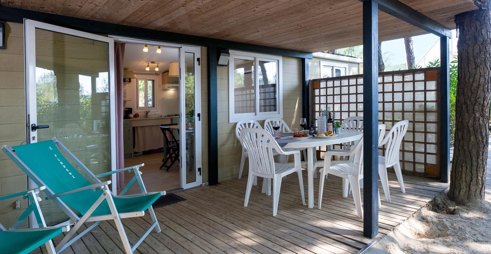 Veranda accogliente con tavolo apparecchiato, sedie bianche e sdraio verdi immerse nella natura.