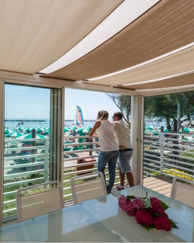 Vista mare da veranda, coppia osserva spiaggia affollata, ombrelloni verdi, atmosfera estiva e rilassata.