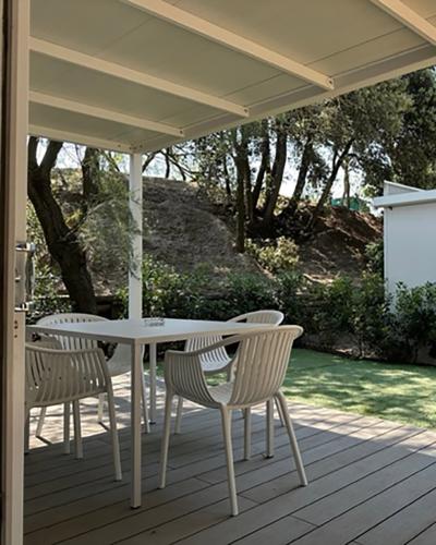 Terrazza accogliente con tavolo bianco, sedie, giardino verde e alberi sotto un cielo sereno.