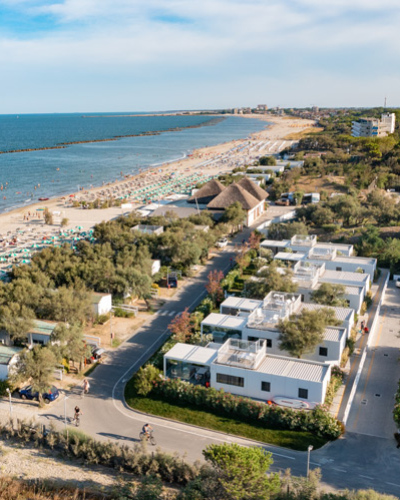 Spiaggia affollata, mare blu, ombrelloni colorati, villette immerse nel verde, estate vivace e rilassante.