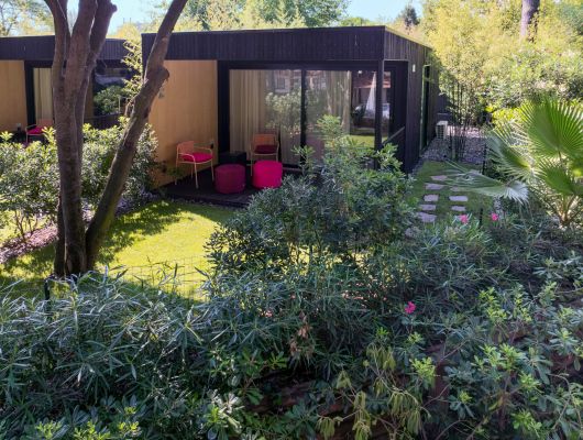 Giardino sereno con bungalow moderno, sedie colorate e natura rigogliosa avvolta dal sole.