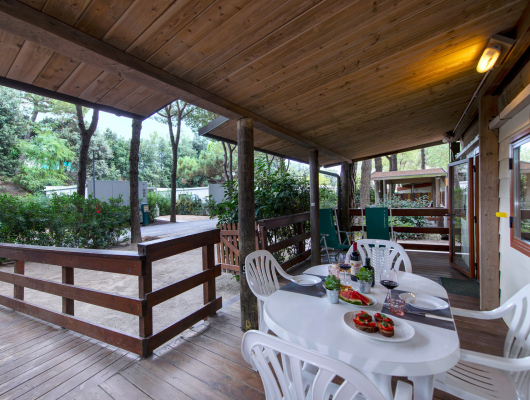 Terrazza in legno tra alberi, tavolo apparecchiato con vino e bruschette, atmosfera rilassante.
