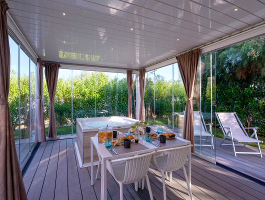 Veranda luminosa con tavolo apparecchiato, vasca idromassaggio e vista su giardino verde rigoglioso.