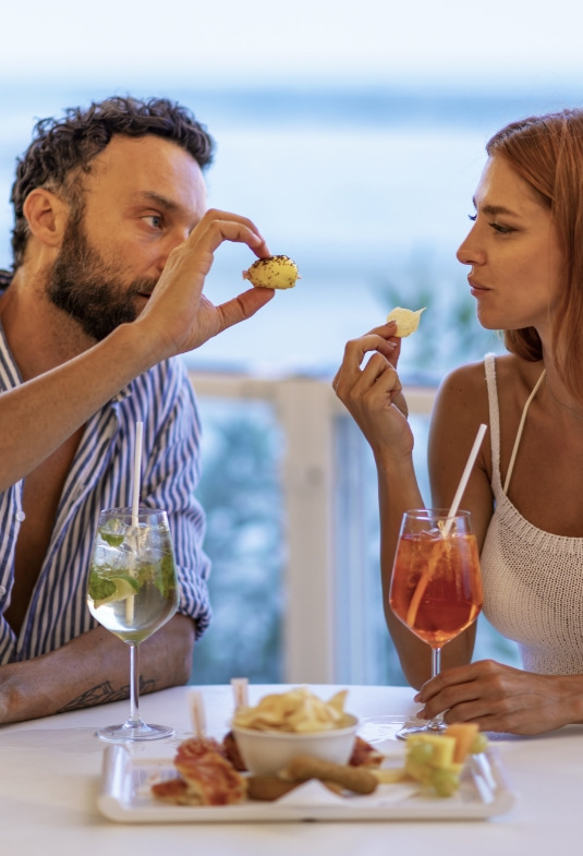 Coppia sorride condividendo cibo e bevande, atmosfera romantica e rilassata al mare.