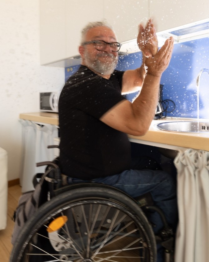 Uomo in sedia a rotelle, felice, spruzza acqua in una cucina accessibile.