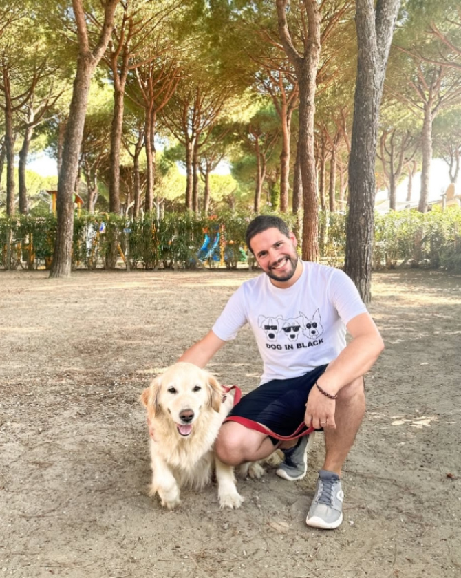 Uomo sorridente accarezza un cane felice in un parco ombreggiato da pini.
