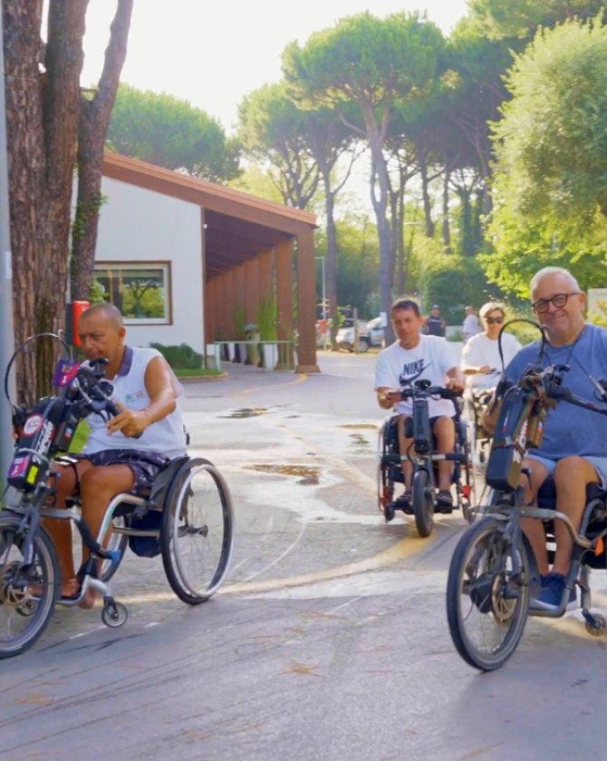 Persone in carrozzina sorridono mentre pedalano all'aperto, circondati da alberi e sole.