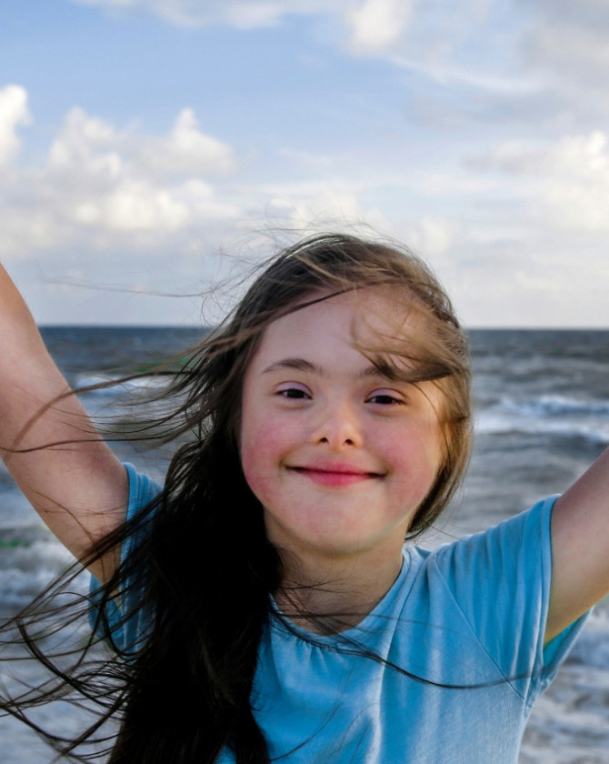 Bambina felice con braccia alzate, sorridente davanti al mare mosso e cielo sereno.