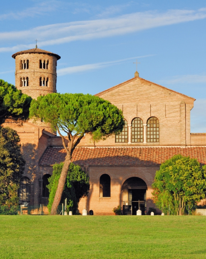 Antica chiesa in mattoni, torre rotonda, pini e cielo sereno evocano pace e storia.