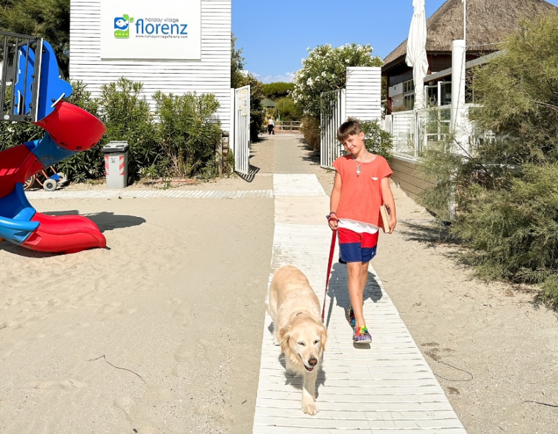 Bambino felice passeggia col cane su passerella, sotto un cielo azzurro, vicino al mare.