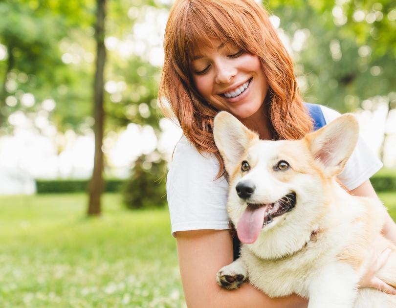 Donna felice abbraccia un cane corgi sorridente in un parco verde e soleggiato.