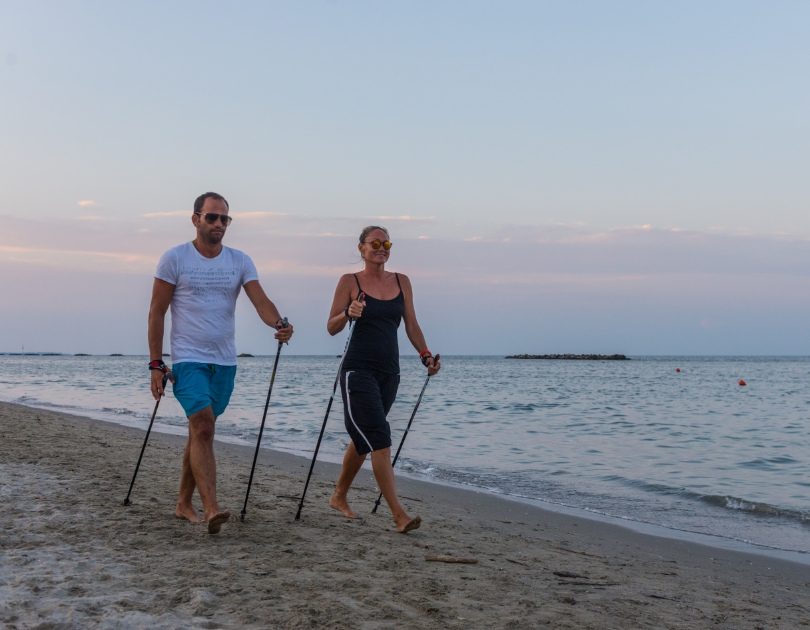 Due persone camminano sulla spiaggia al tramonto, usando bastoncini, aria serena e rilassata.